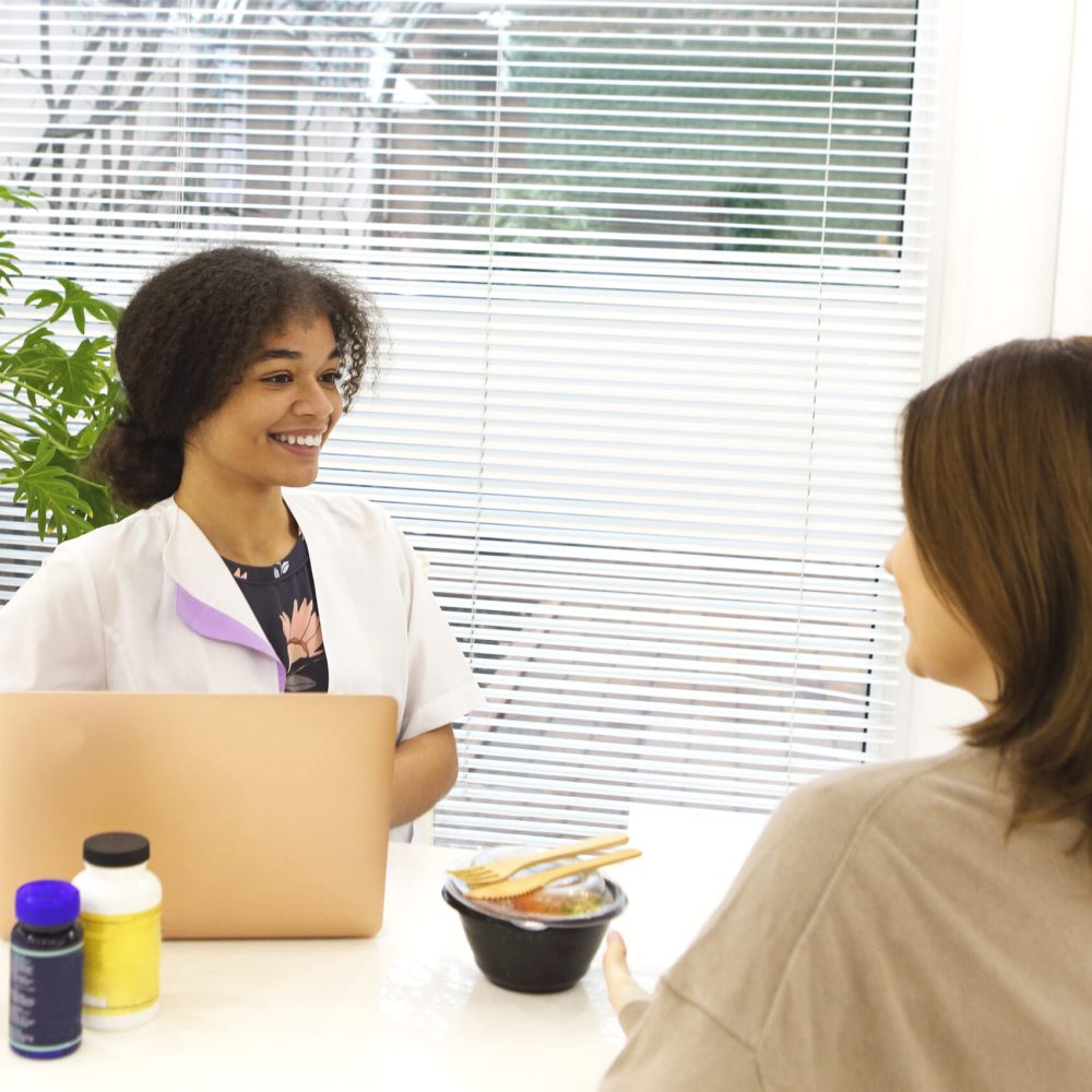 Smiling black lady doctor chatting with female patient at clinic advicing her vitamins and fresh takeaway salad in a lunch box. Dietology and nutririon concept